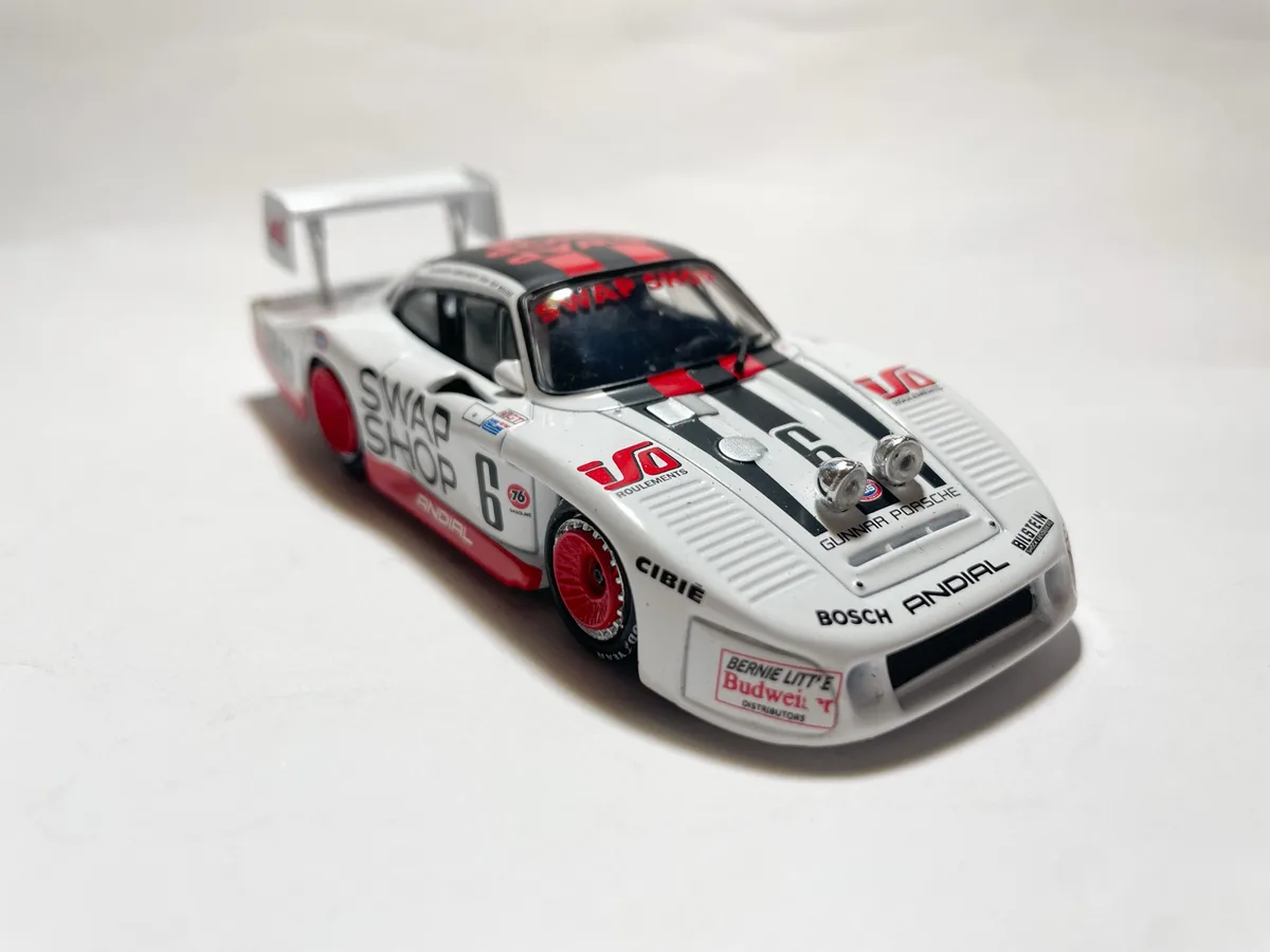 A white and red die-cast model of a Porsche race car with black racing stripes, featuring sponsor decals like 'SWAP SHOP'…
