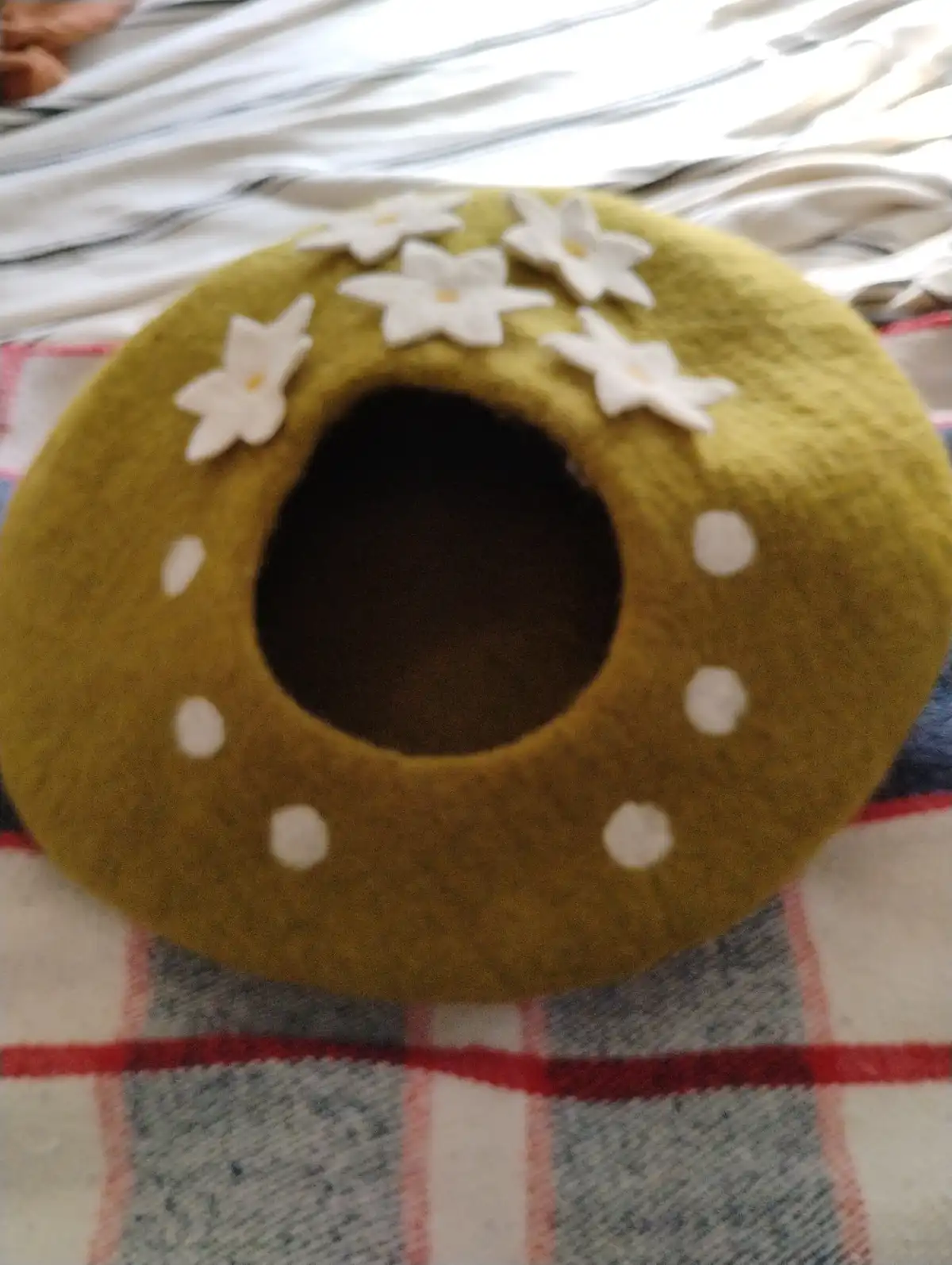 A handmade, olive-green felt pet bed shaped like a donut with a large central hole, decorated with white felt flowers and…