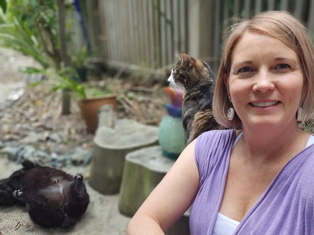 A woman smiles at the camera while a tabby cat sits calmly on her shoulder, looking away. In the foreground, a black cat…