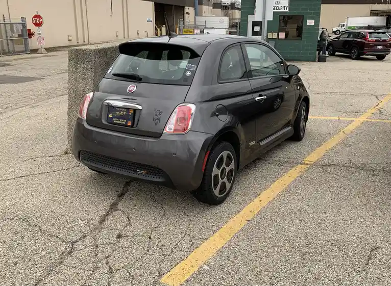 A dark gray Fiat 500e electric car is parked in a lot, angled slightly into a concrete barrier. The car has a Michigan…