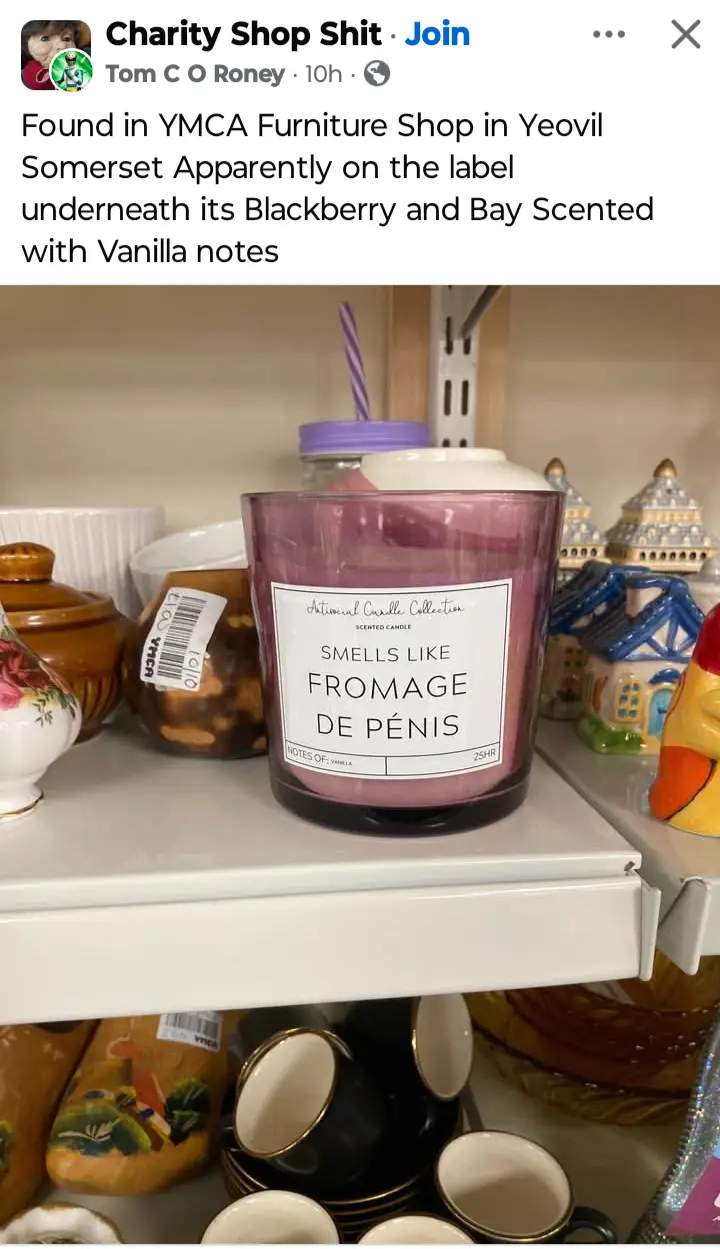 A pink scented candle in a glass jar sits on a shelf in what appears to be a charity shop, surrounded by various ceramic and…