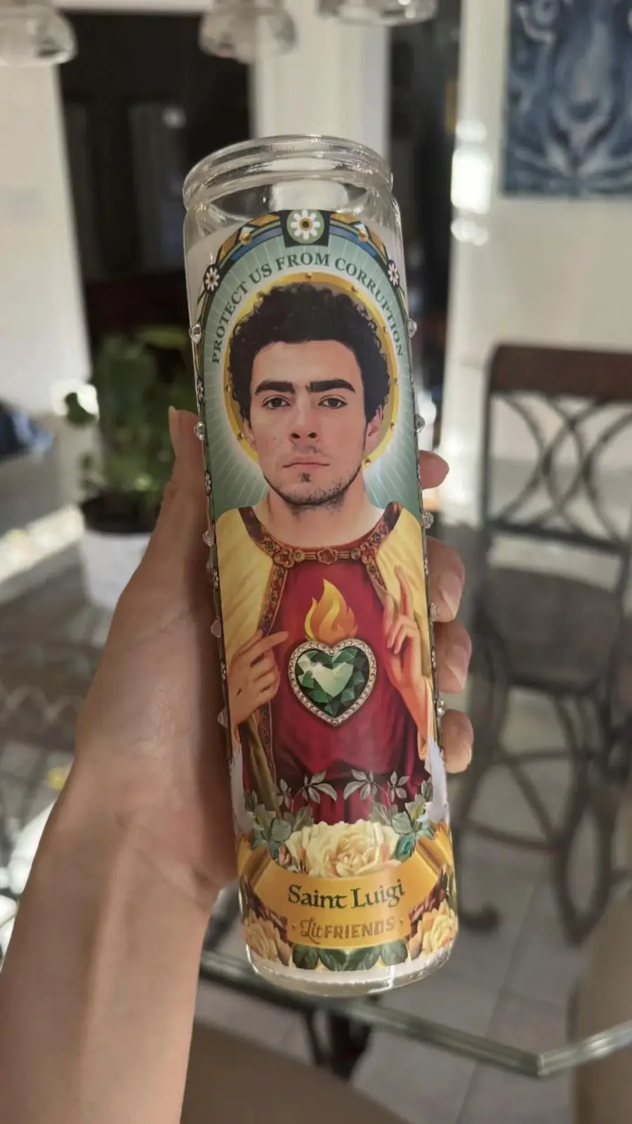 A hand holds a decorative prayer candle featuring a saintly portrait of a man with dark hair and a goatee, wearing a red…