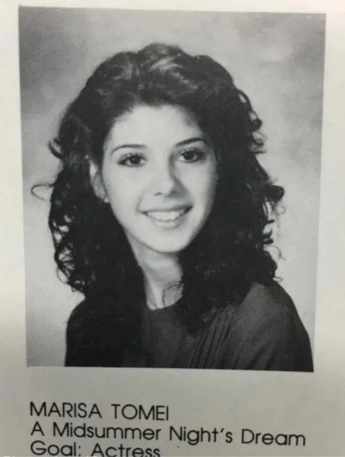 A black-and-white school yearbook photo of a young woman with curly dark hair and a smile, wearing a dark top. Below the…