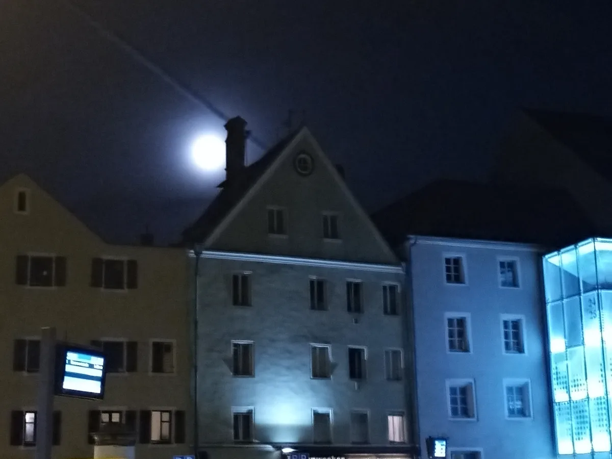 A nighttime photo of several European-style buildings with lit windows under a dark sky, where a bright full moon is visible…