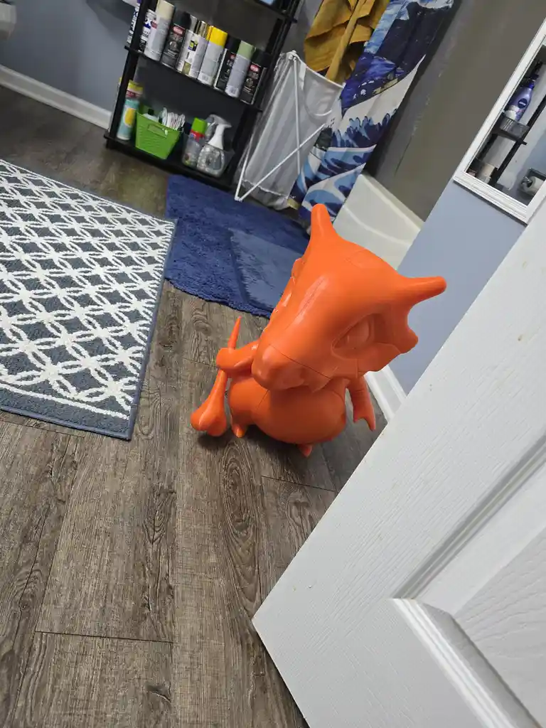 An orange plastic dragon figurine sits on a wooden floor in a bathroom, positioned near a white door and next to a blue rug…