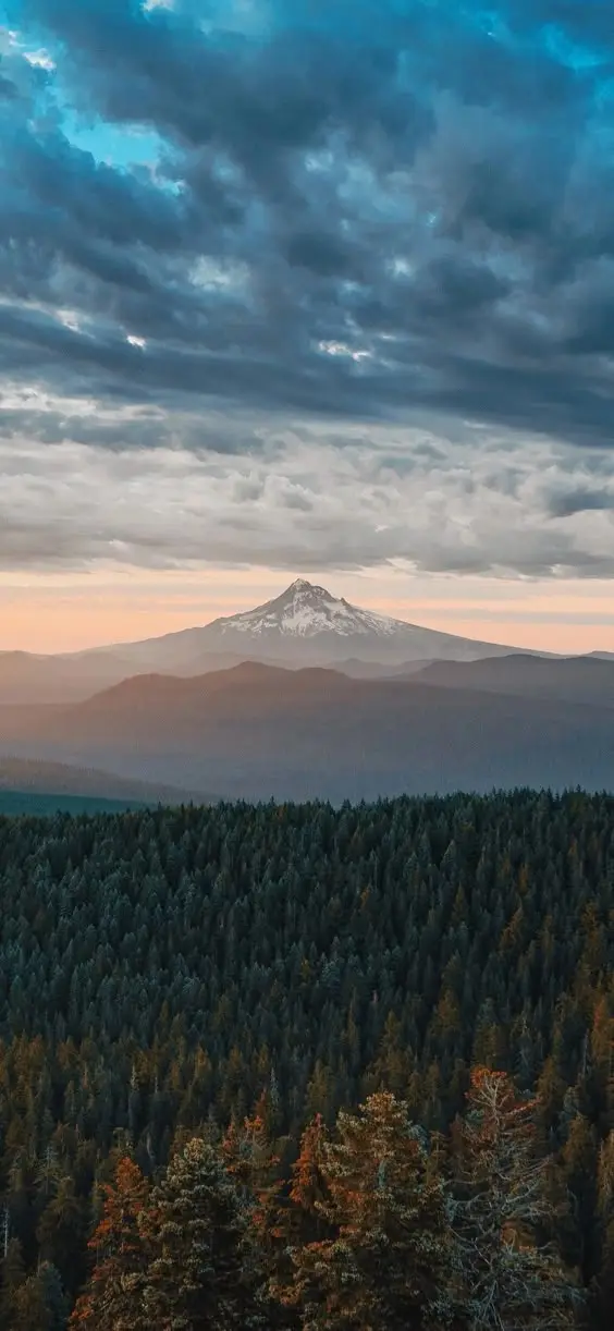 A scenic landscape photo shows a snow-capped mountain peak under a dramatic cloudy sky, with layers of rolling hills and a…