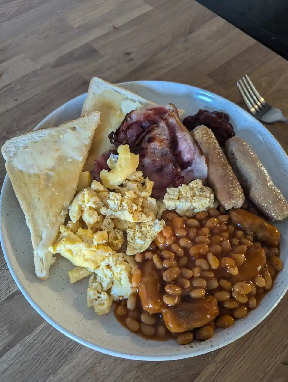 A plate of a full English breakfast with scrambled eggs, baked beans, bacon, sausages, buttered toast, and a small portion…