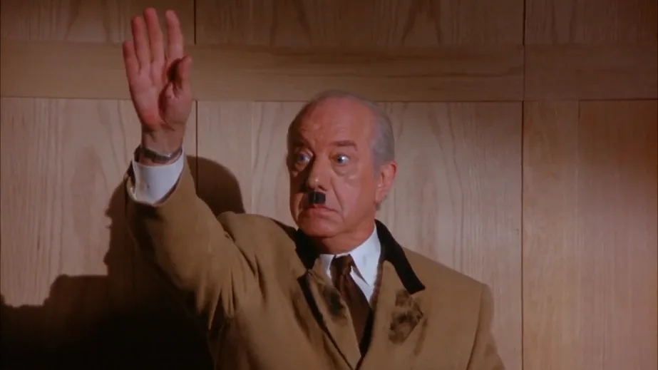 a man in a brown suit with a mustache raises his hand in a salute while looking shocked, standing against a wood-paneled…