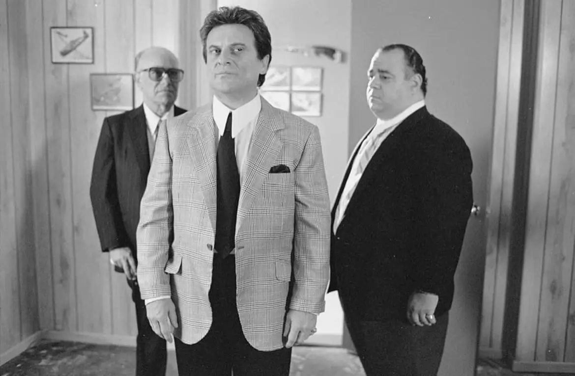A black-and-white photo shows three men in suits standing in a hallway. The man in the center, wearing a plaid blazer and…