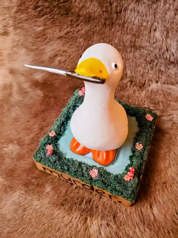 A small white rubber duck with orange feet sits in a miniature pond made of green moss and blue resin, decorated with tiny…