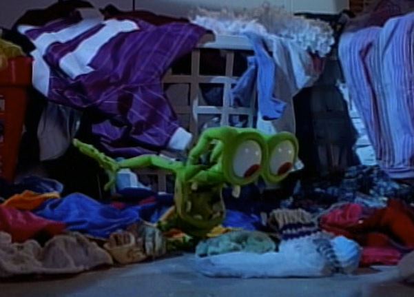 A green, cartoonish frog-like creature with large, bulging eyes sits amidst a chaotic pile of colorful laundry on the floor…