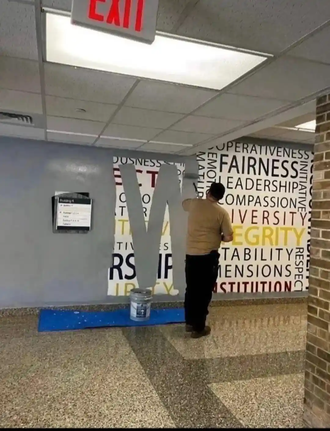 A person is painting over a wall mural filled with positive words like 'fairness', 'leadership', 'compassion', and…