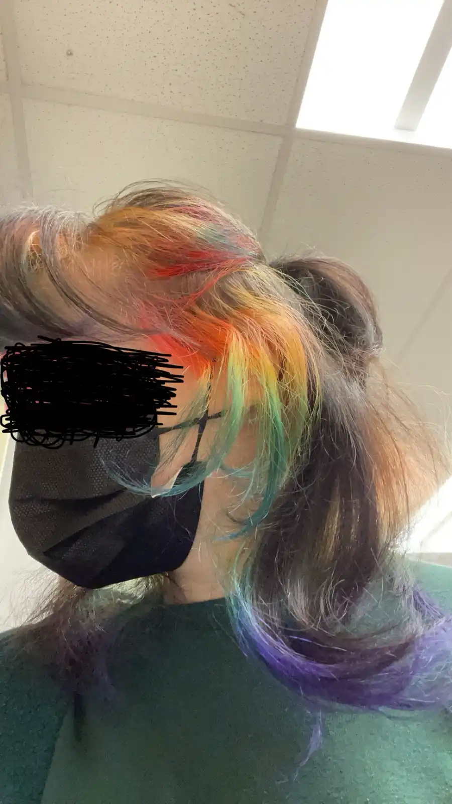 A person wearing a black face mask with their face obscured by black scribbles, showing rainbow-colored hair in a ponytail…