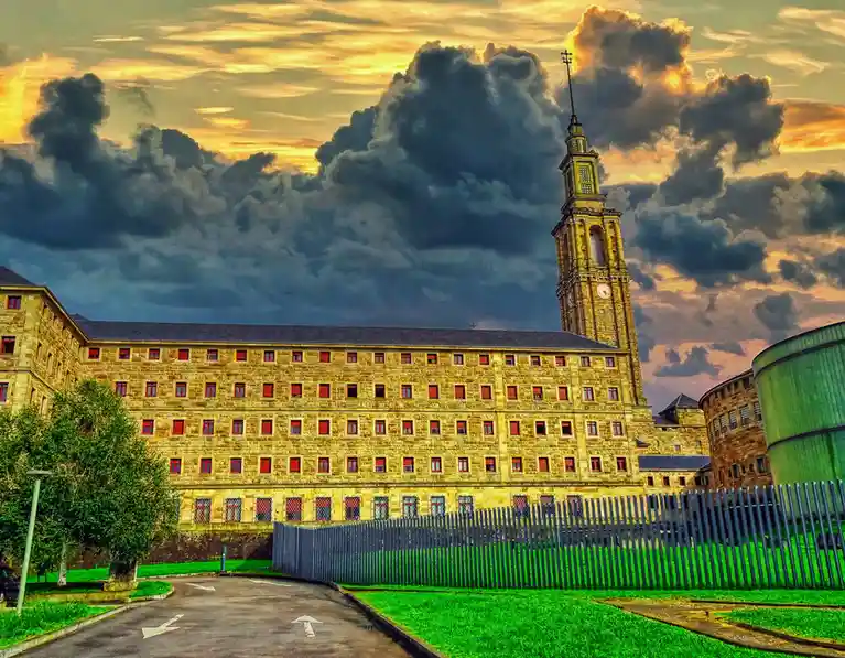 A grand, stone building with a tall clock tower and many windows, set against a dramatic sunset sky with dark clouds. A…