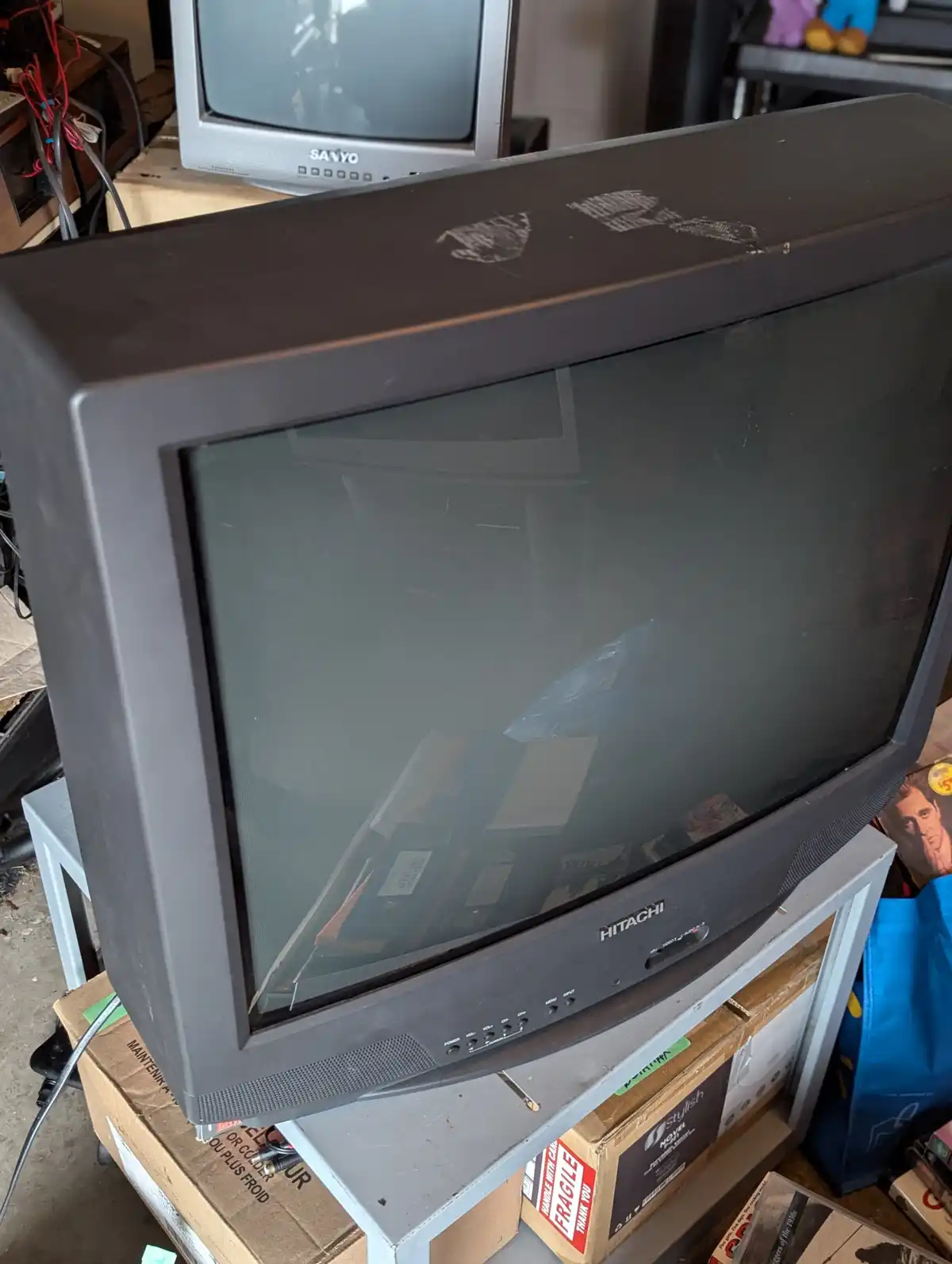 A dusty, old Hitachi CRT television sits on a shelf surrounded by boxes and clutter, with the brand name visible on the…
