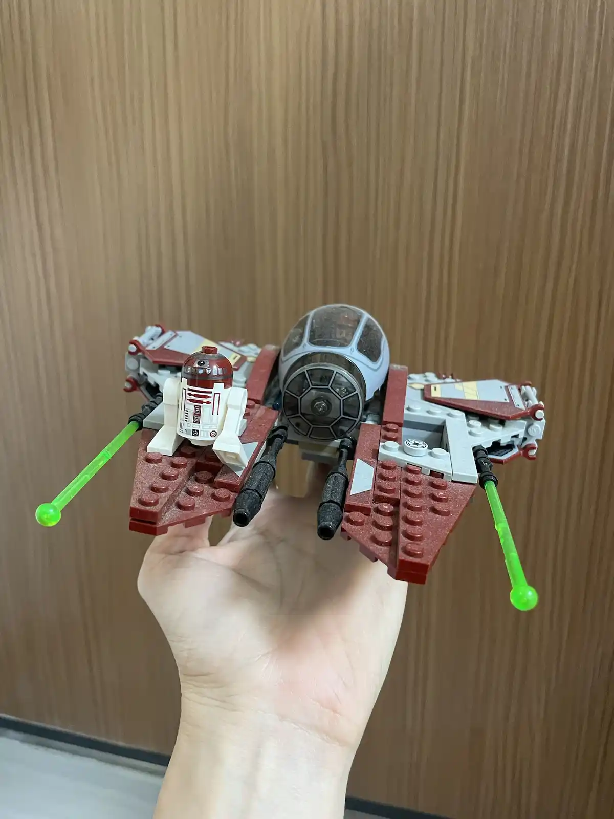 A hand holds a LEGO Star Wars starfighter model, predominantly red and gray, with a small R2-D2 minifigure seated in the…
