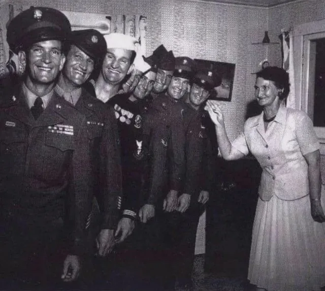 A black-and-white photo shows a woman in a light-colored dress smiling and waving at a group of smiling men in military…
