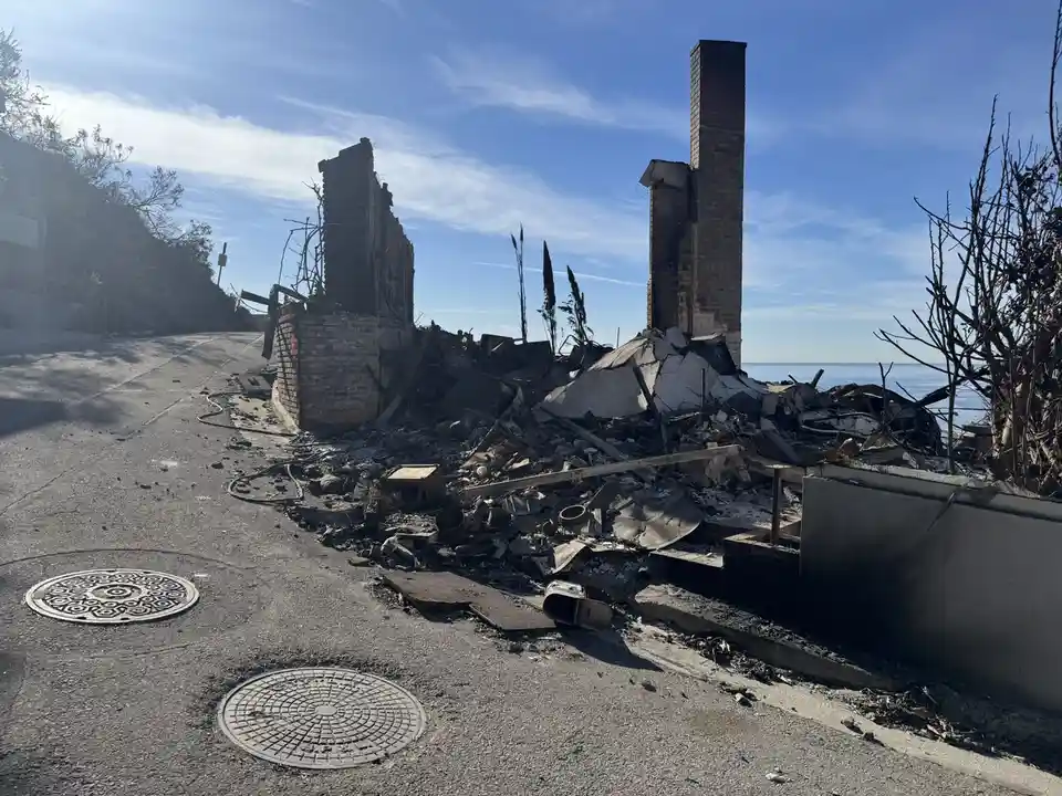 A photograph shows the charred remains of a house next to a road, with only a brick chimney and wall standing amidst rubble…