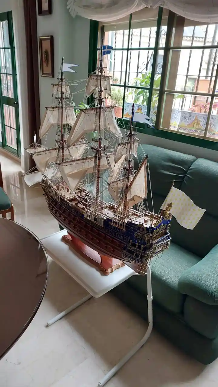 A highly detailed model of a multi-masted sailing ship with beige sails and blue/gold accents sits on a white stand in a…