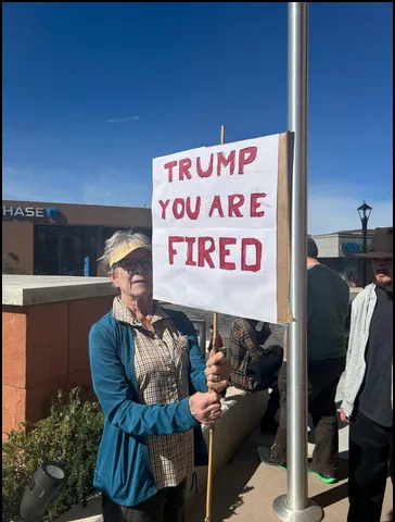 An older woman with a yellow headband and blue jacket holds a handmade white sign with red handwritten text that reads…