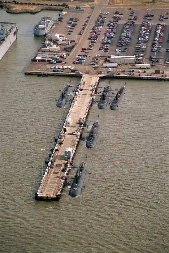 An aerial view shows several submarines docked in a line along a pier, with a large parking lot full of cars visible on land…