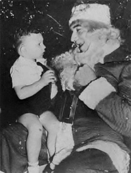 A grainy black-and-white photo shows a smiling man dressed as Santa Claus holding a small child on his lap. The child looks…
