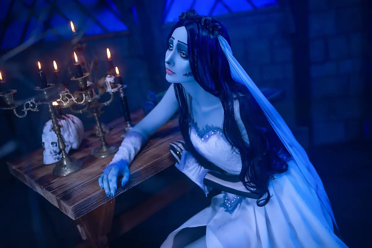 A person in a white wedding dress and veil with dark makeup and long black hair sits at a wooden table in a dimly lit…