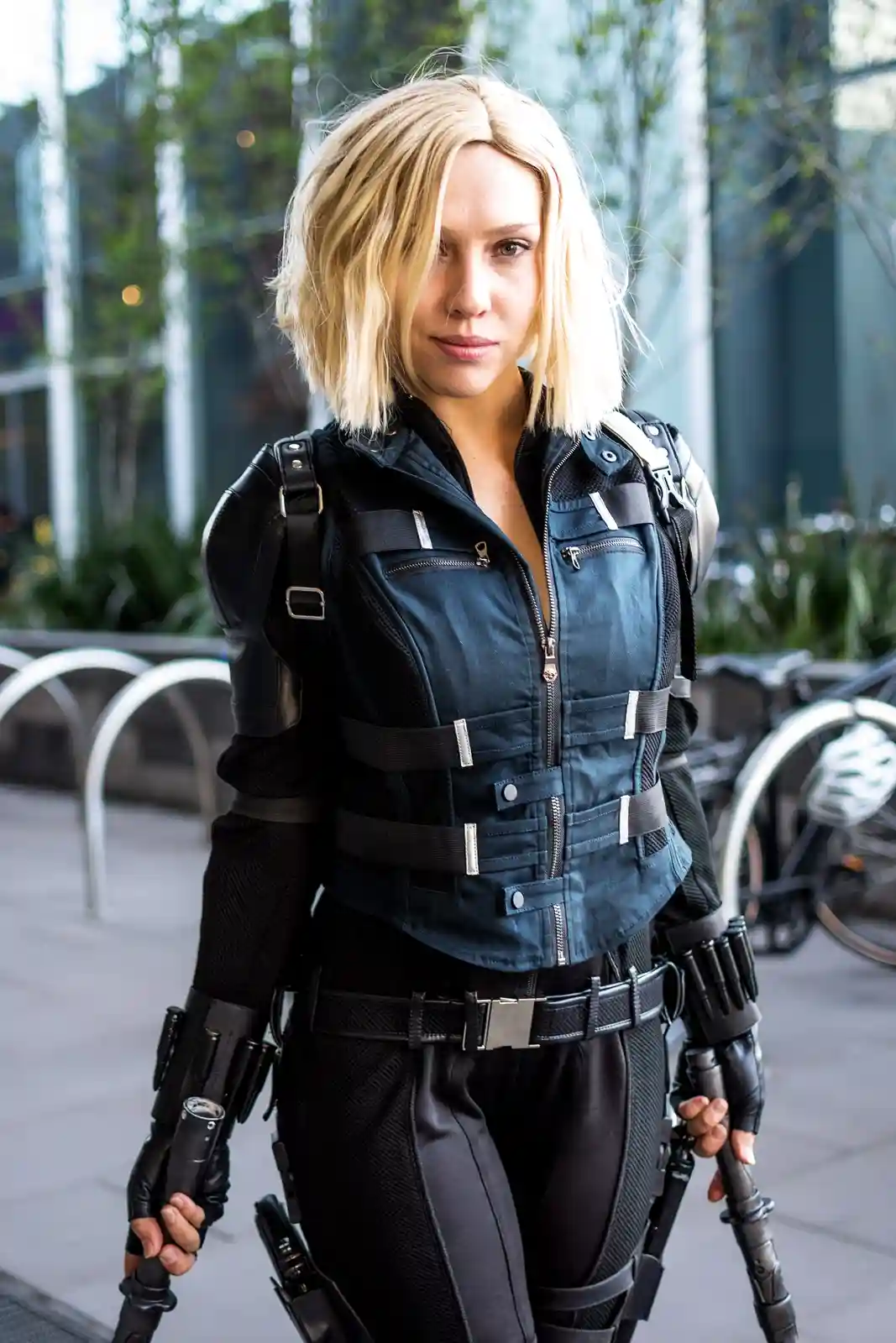 A woman with short blonde hair wears a dark tactical outfit with straps, zippers, and utility gear, holding weapons. She…