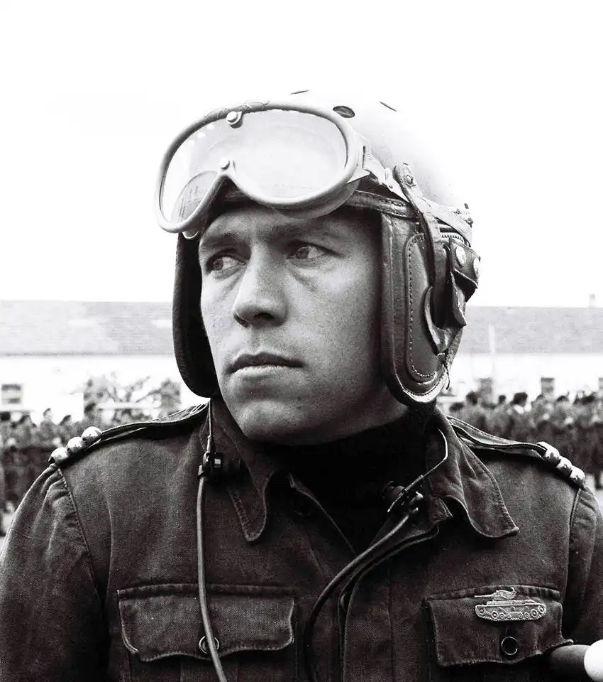 A black-and-white photo shows a man in a military flight helmet with goggles pushed up, looking off to the side with a…