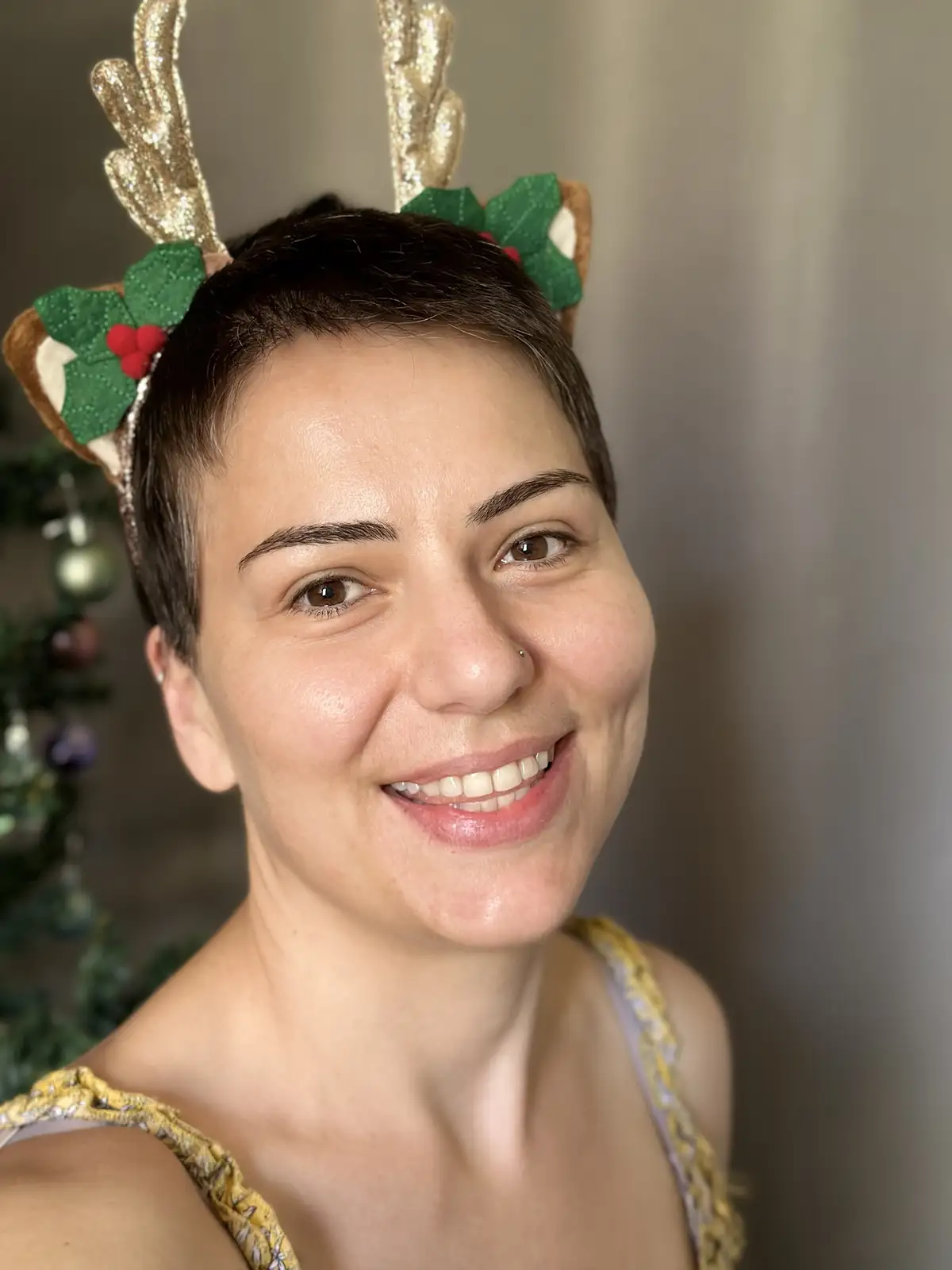 A person with short dark hair smiles at the camera while wearing a festive headband with glittery gold reindeer antlers and…