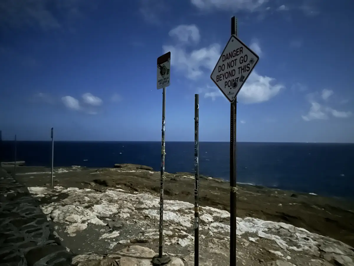 A photo of a rocky cliffside overlooking the ocean with two warning signs on poles. The prominent diamond-shaped sign reads…