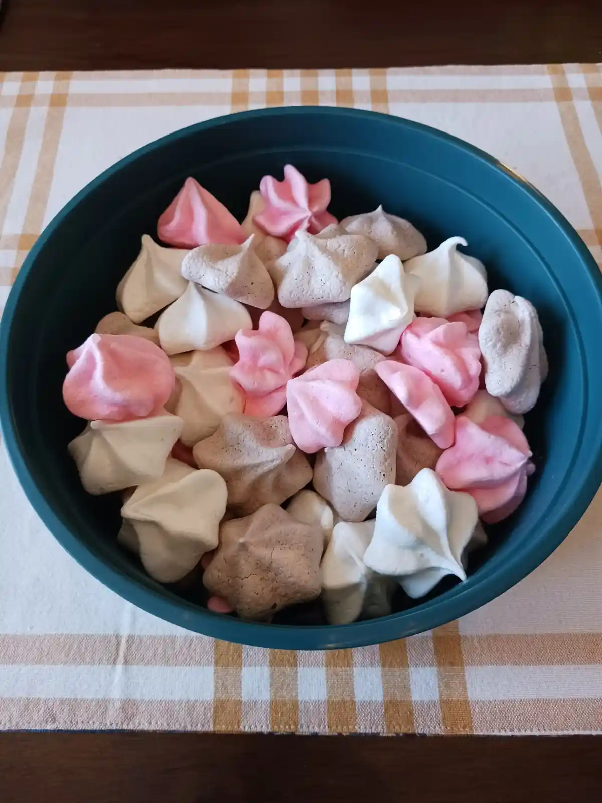 A teal bowl filled with homemade meringues in pink, white, and beige colors sits on a checkered tablecloth. The meringues…