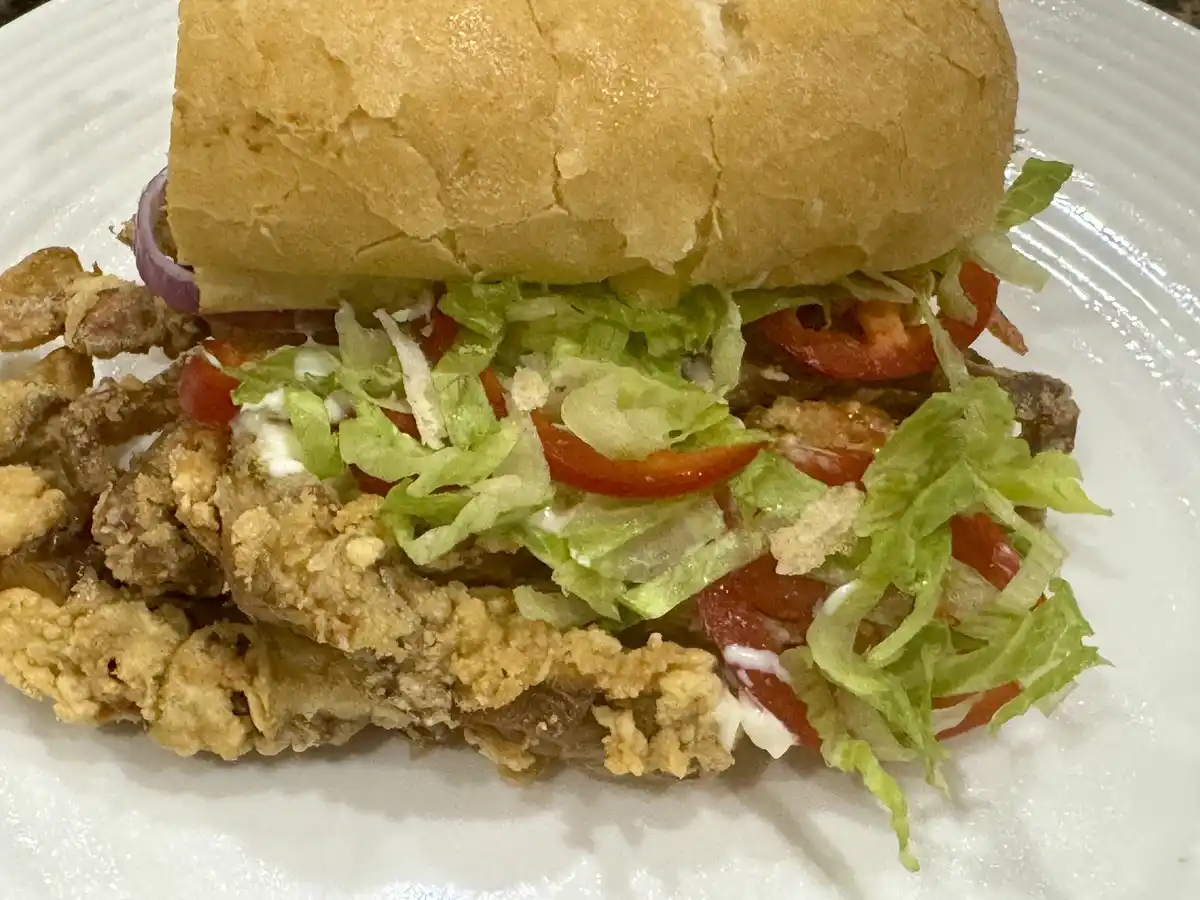 A close-up photo of a sandwich on a white plate, featuring a golden-brown bun, crispy fried meat, shredded lettuce, sliced…