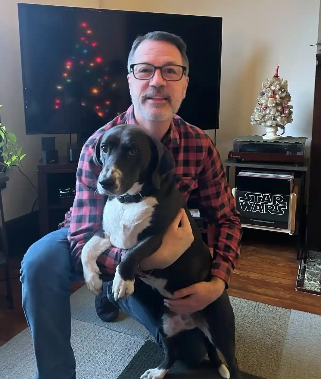 A man with glasses and a red plaid shirt is kneeling and holding a black and white dog in his lap. Behind him, a TV displays…