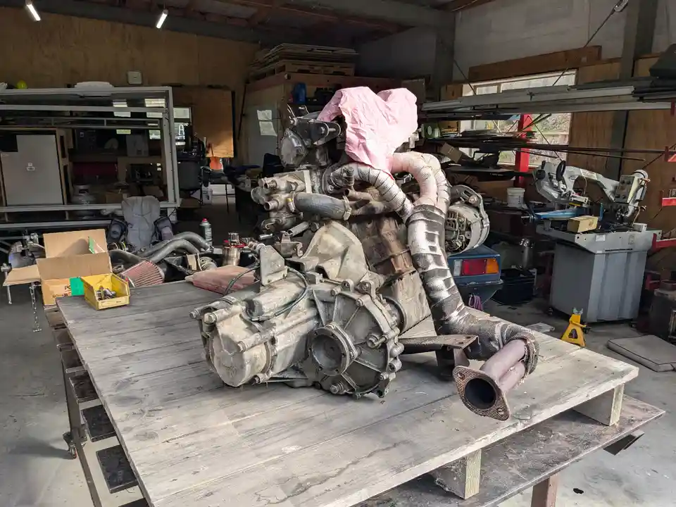 A large, dirty car engine sits on a wooden workbench in a cluttered garage, with various tools and parts scattered around. A…