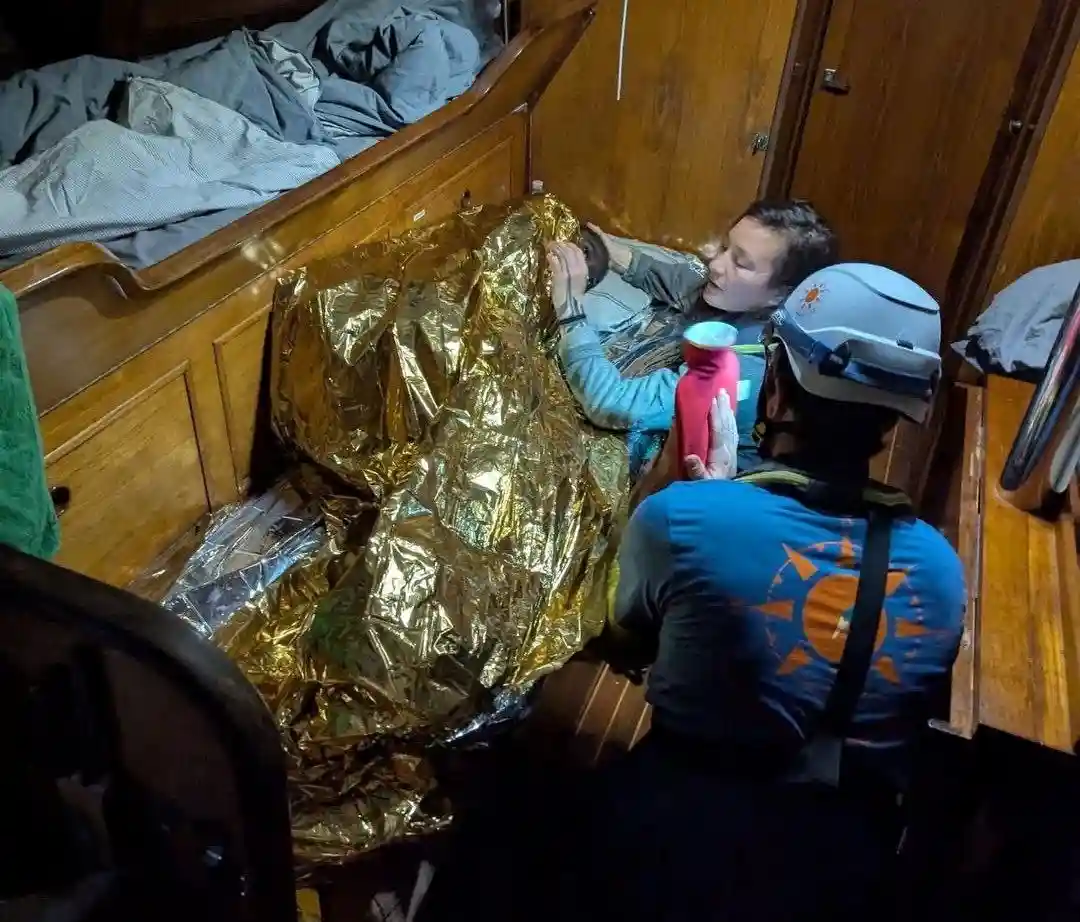 A person in a blue shirt and white helmet sits beside a bed on a boat, holding a pink water bottle while another person lies…