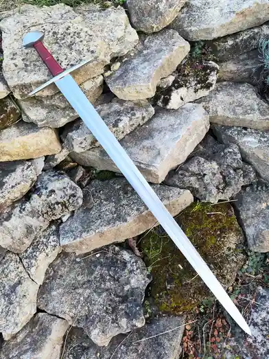 A plastic sword with a red grip and silver guard rests diagonally across a pile of rough, mossy stones. The blade is…