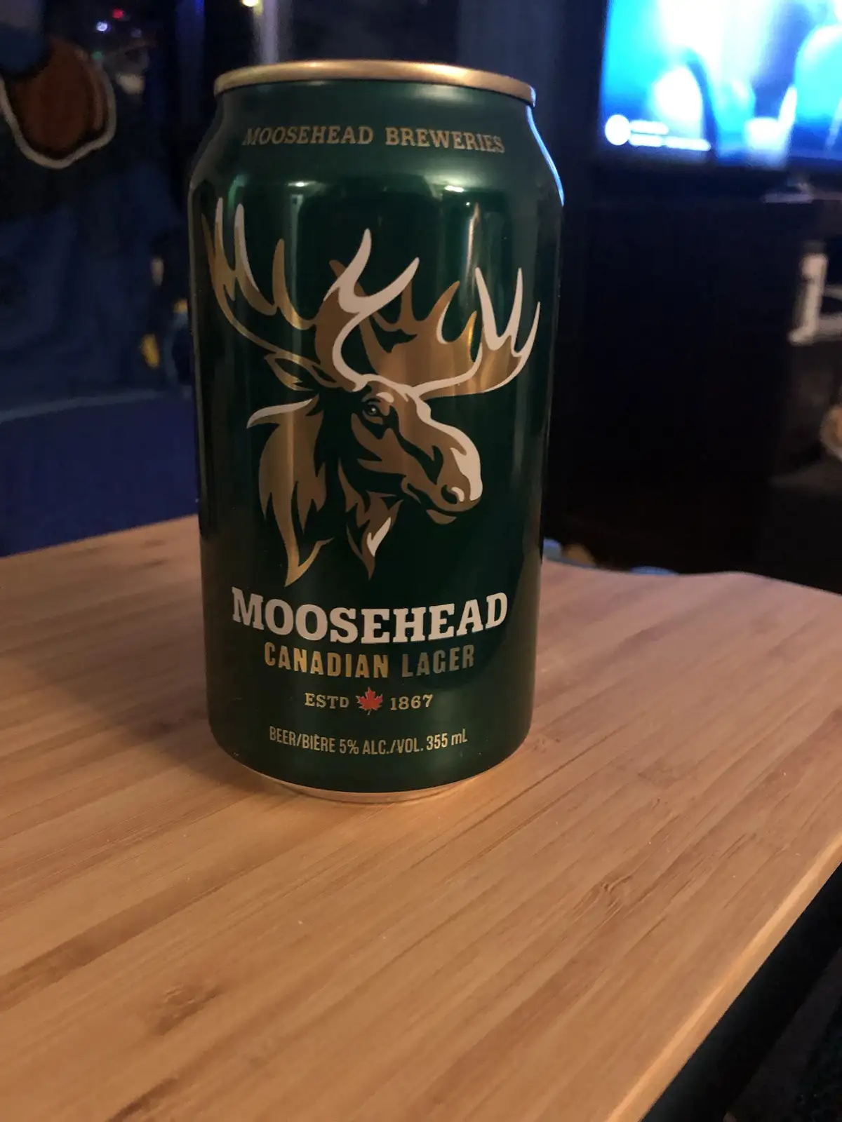 A green can of Moosehead Canadian Lager beer, featuring a large moose head logo, sits on a wooden table. The can displays…