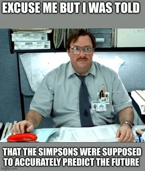 A man with glasses and a mustache sits at a cluttered office desk, wearing a gray shirt and tie with a name badge. He has a…