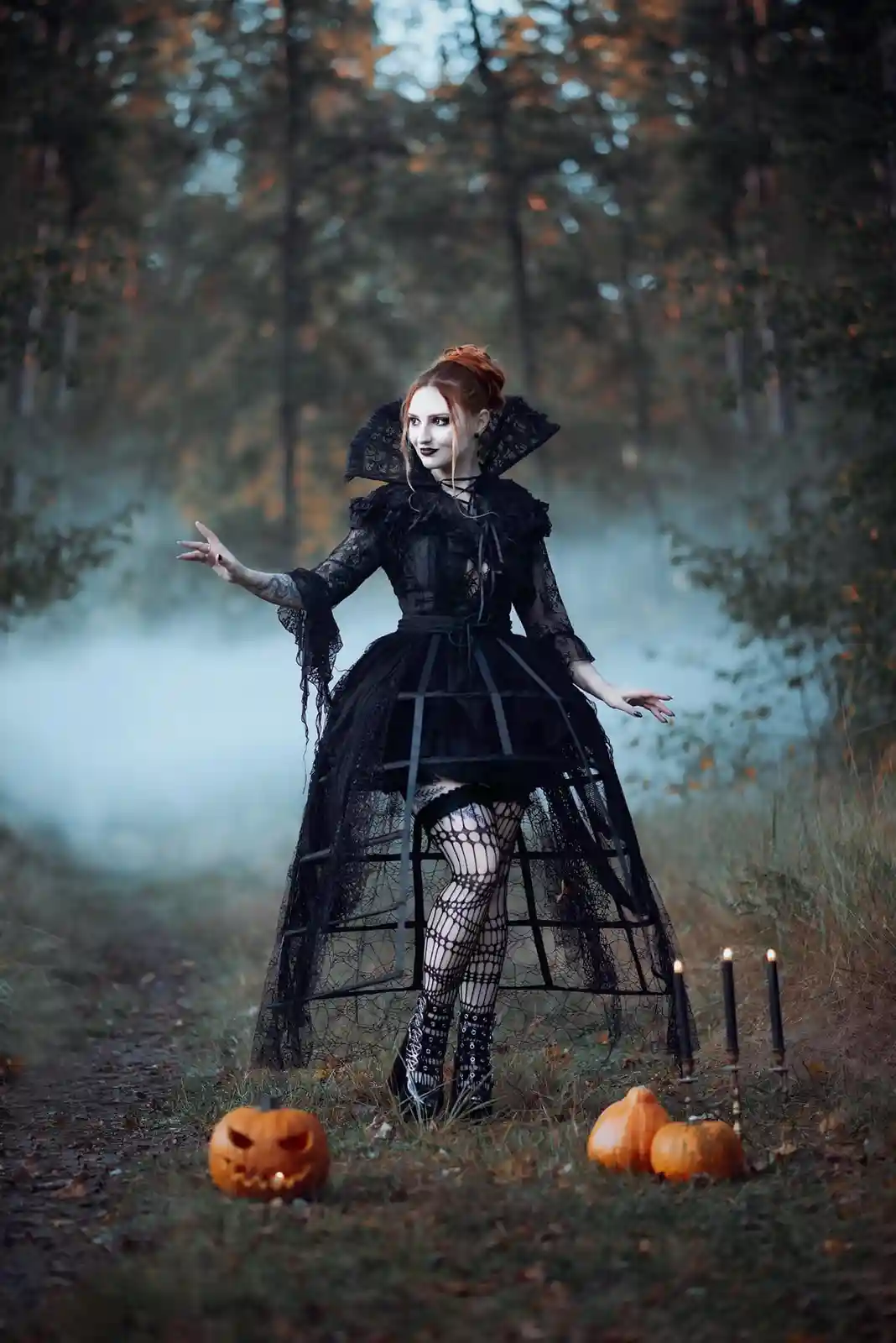 A woman in a black gothic witch costume with lace, a high collar, and fishnet stockings stands in a misty forest path. She…