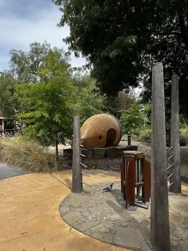 A photograph of a park playground with a large wooden acorn-shaped structure, wooden posts with metal bars, and a stone…