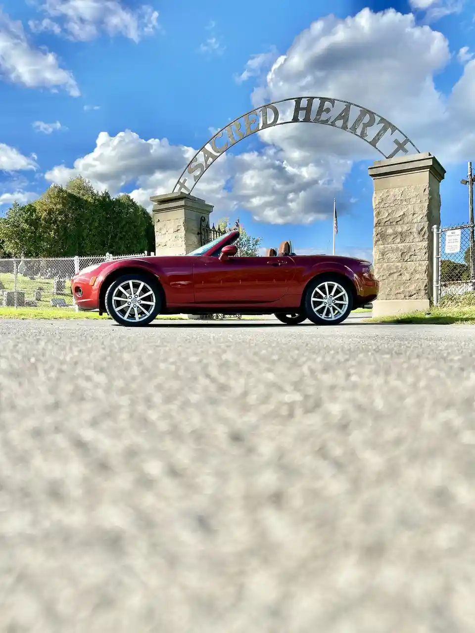 A red convertible sports car is parked in front of a large metal archway that reads 'SACRED HEART' flanked by stone pillars…