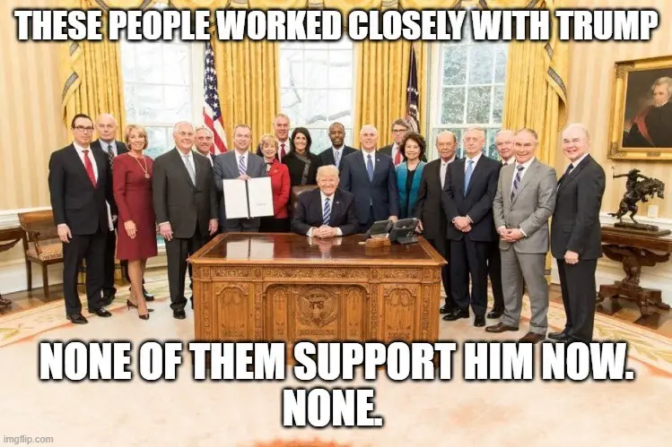 A group photo of numerous people, mostly in business attire, standing and sitting around a large wooden presidential desk in…