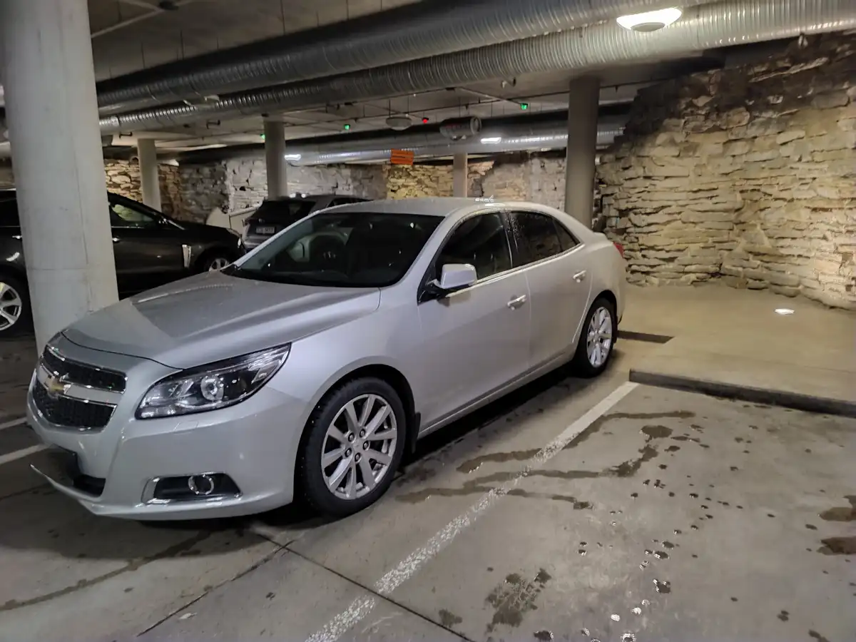 A silver Chevrolet sedan is parked in an underground parking garage with wet concrete and stone walls. The car's surface is…