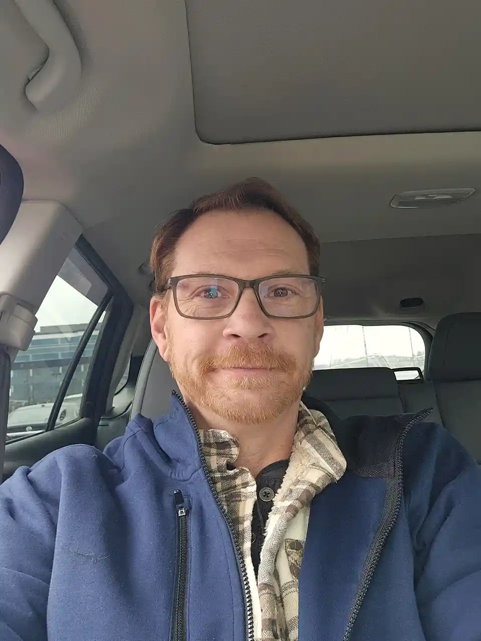 A man with glasses and a reddish beard takes a selfie inside a car, wearing a blue zip-up hoodie over a plaid shirt. The…