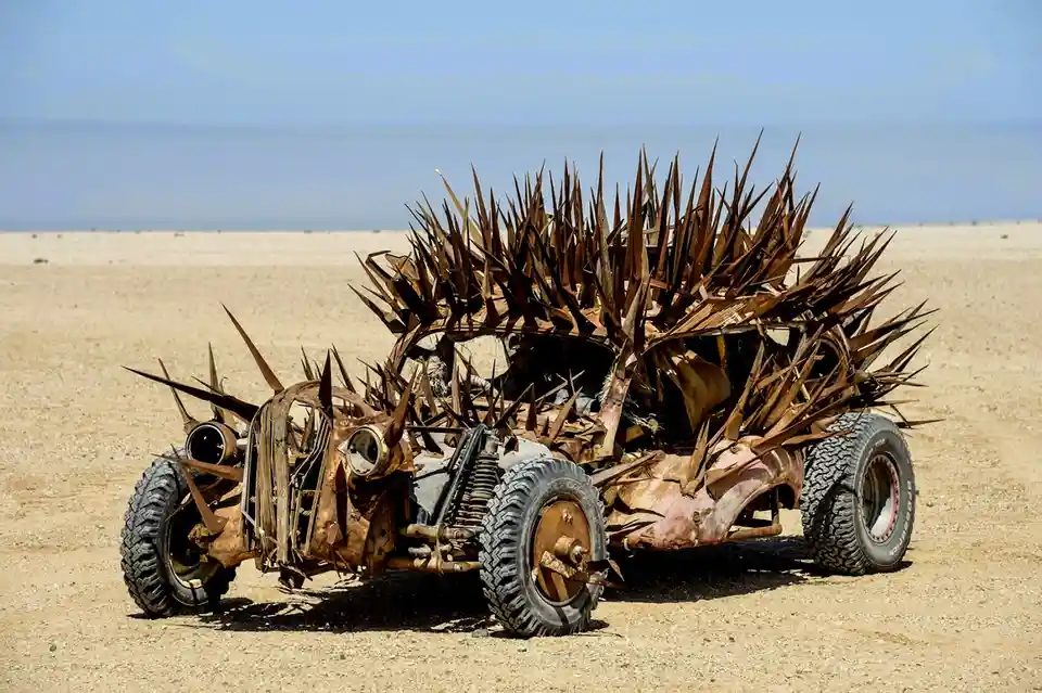 A heavily modified, rusted, and spiked vehicle resembling a hedgehog stands in a barren desert landscape under a clear blue…