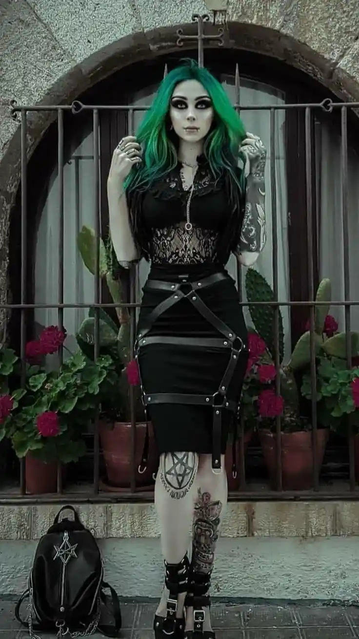 A person with vibrant green hair, heavy black makeup, and multiple tattoos stands in front of a barred arched doorway. They…