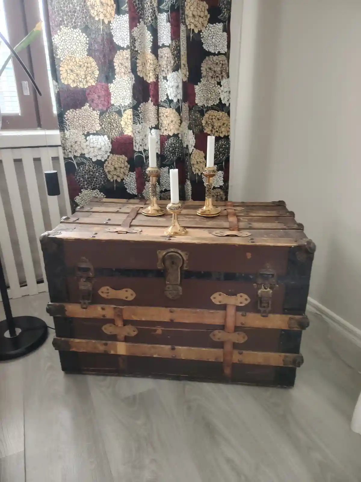An old, weathered wooden trunk with metal latches and leather straps sits on a light wood floor, topped with three ornate…