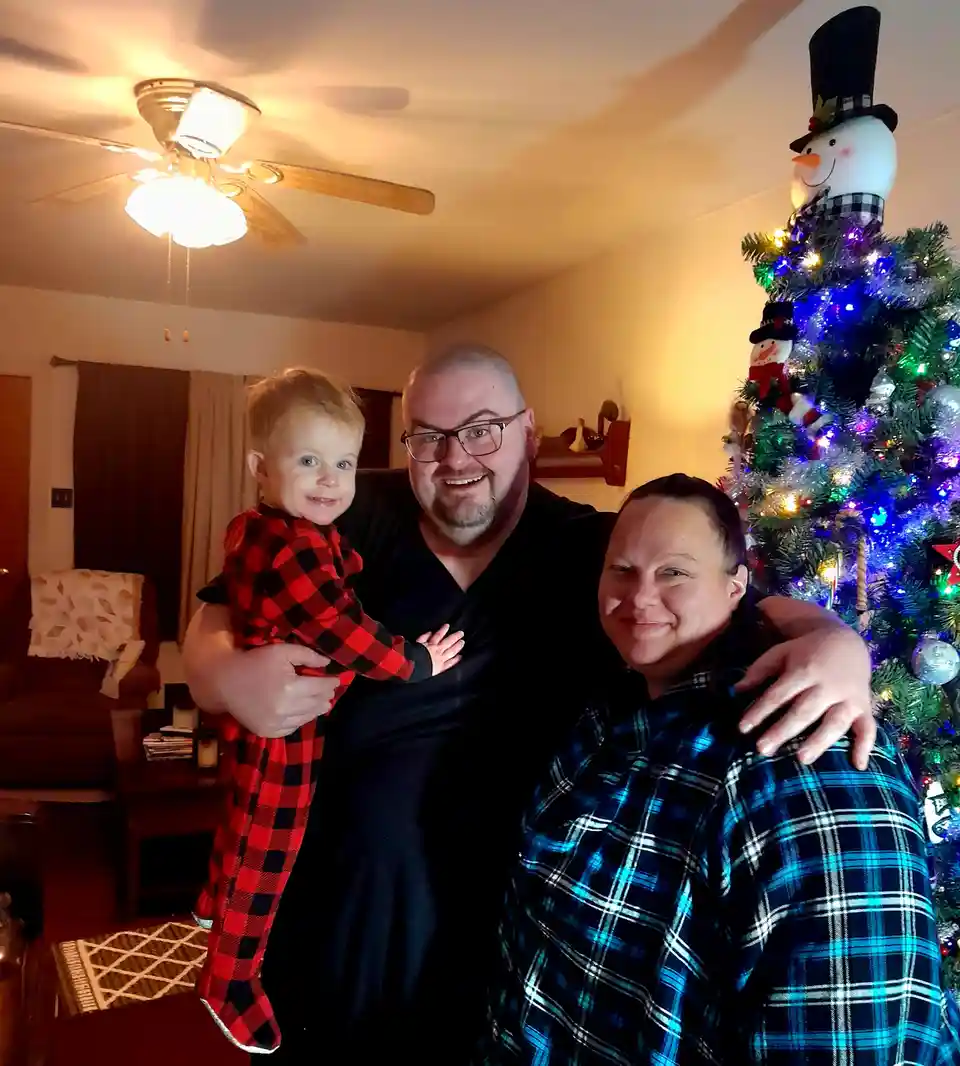 A family photo shows a man holding a toddler in red plaid pajamas while standing beside a woman in blue plaid pajamas, all…