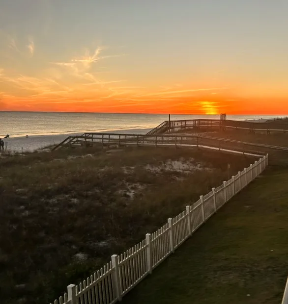 A scenic beach sunset with orange and yellow hues over the ocean, a wooden boardwalk leading to the shore, and a white…